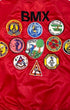 A close-up of a red satin bomber jacket showing the back, decorated with the black embroidered text 'BMX' and a collection of colorful BMX-themed patches.