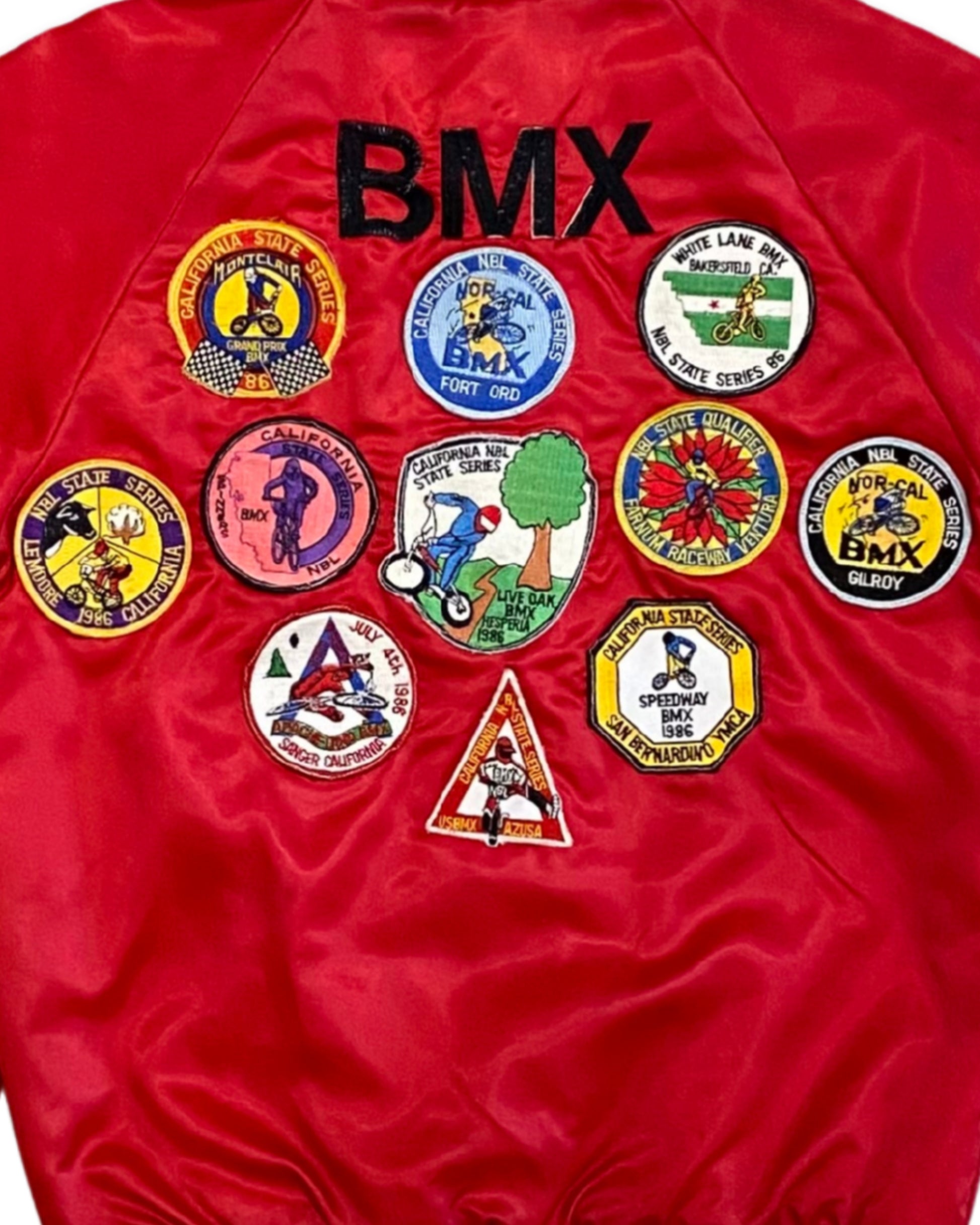 A close-up of a red satin bomber jacket showing the back, decorated with the black embroidered text 'BMX' and a collection of colorful BMX-themed patches.