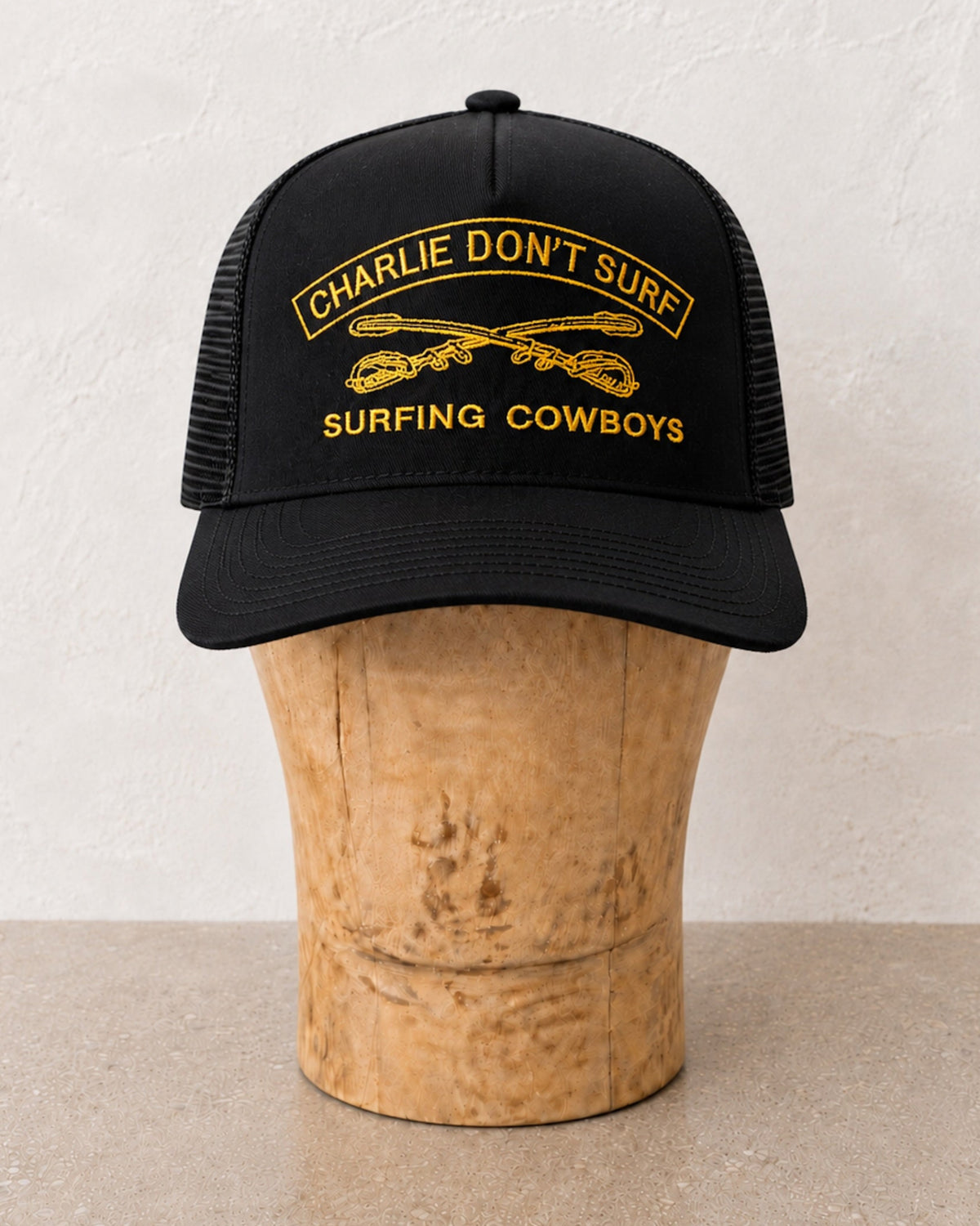 Front view of Charelie Don't Surf Surfing Cowboys embroidered Trucker Cap in Black with Gold - on light wood stand.