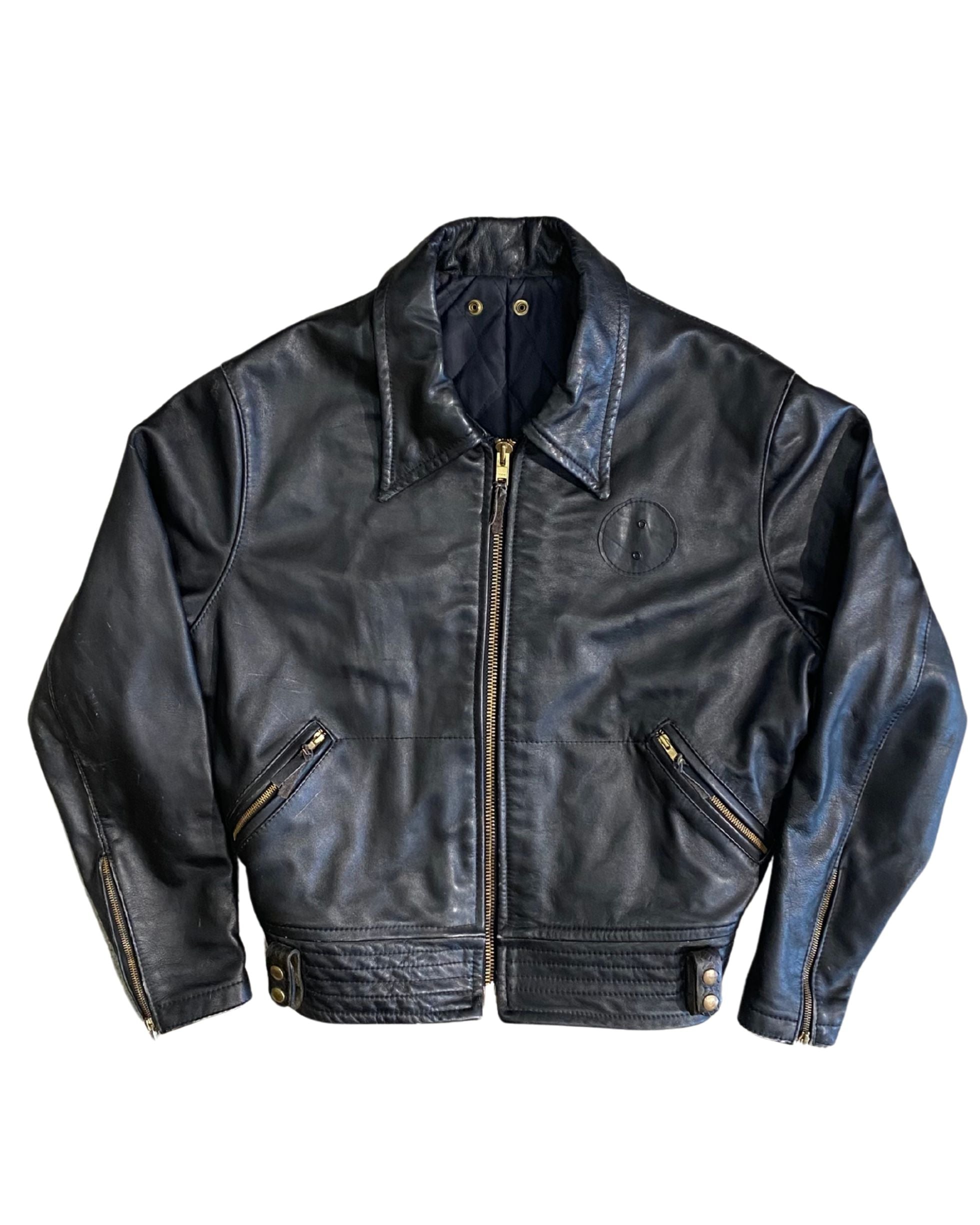 Front view of vintage 1970s black leather motorcycle jacket with brass center zip, angled zip pockets, circular chest medallion, and snap quilted collar, on white background.