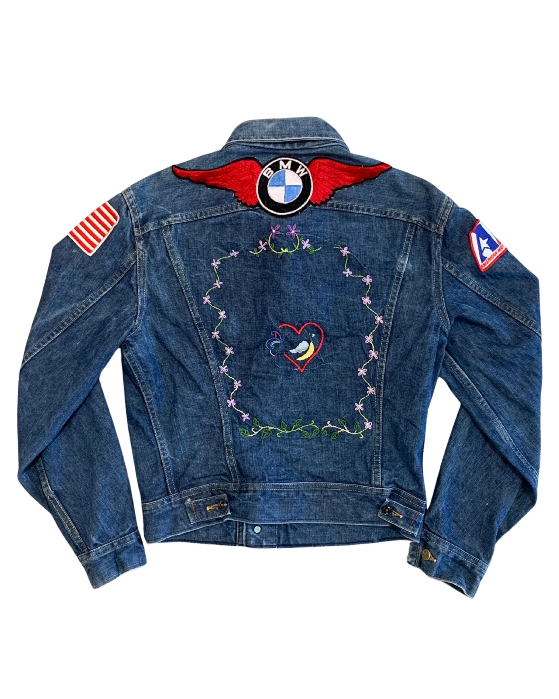 Back view of a faded blue Lee denim jacket with a winged BMW patch, floral embroidery, heart motif, American flag patch, and AMA patch.