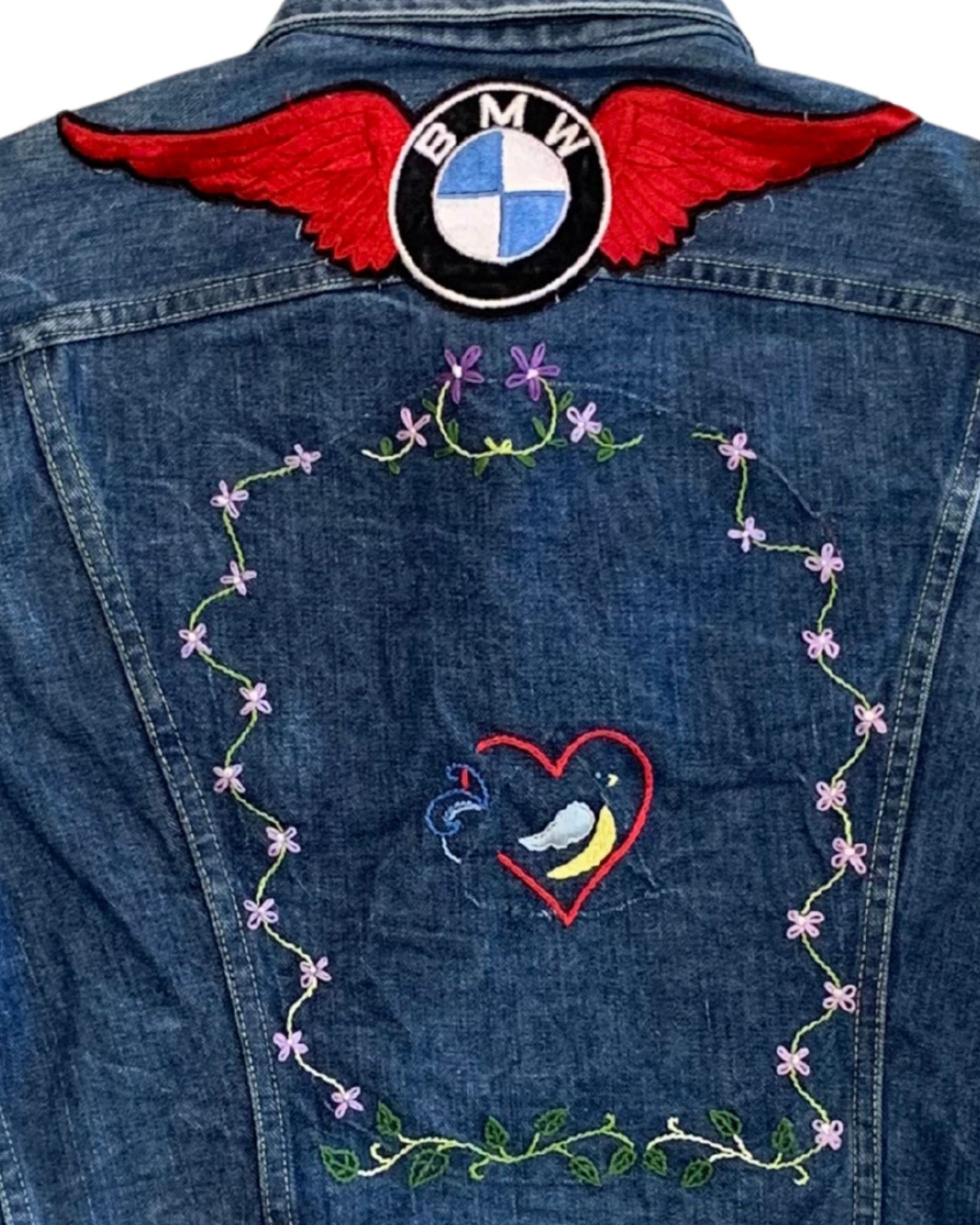 Close-up of the back of a faded denim jacket with a large red-winged BMW patch and hand-embroidered flowers surrounding a small heart motif.