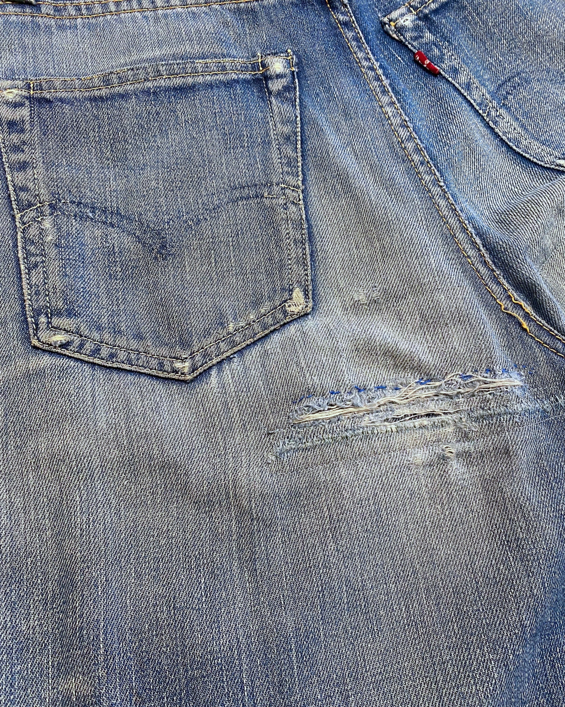 Close-up of the rear seat showing worn back pockets, faded denim, abrasions, tears, and repaired areas below the pockets.