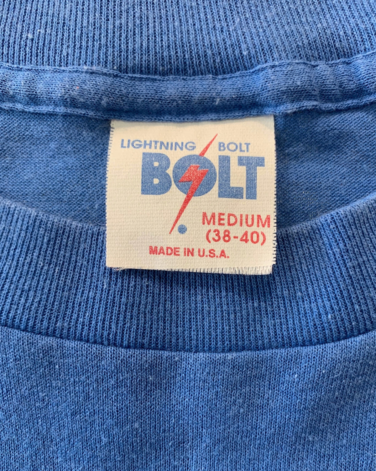 Close-up of the original 1970s woven neck label reading Lightning Bolt Medium 38-40 Made in U.S.A. on a heather blue cotton knit shirt.
