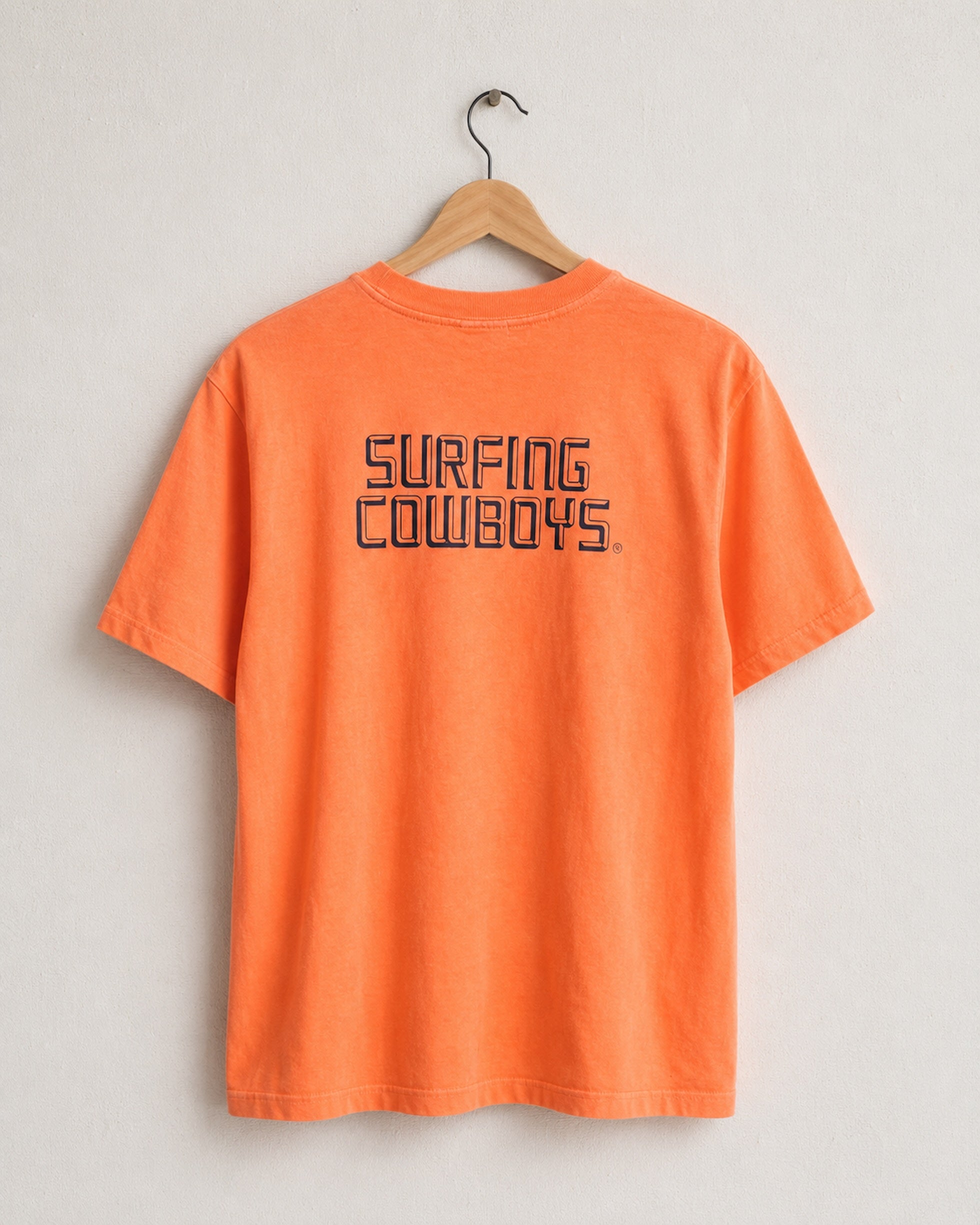 Faded orange t-shirt with 'Surfing Cowboys' text on a hanger against a light gray background