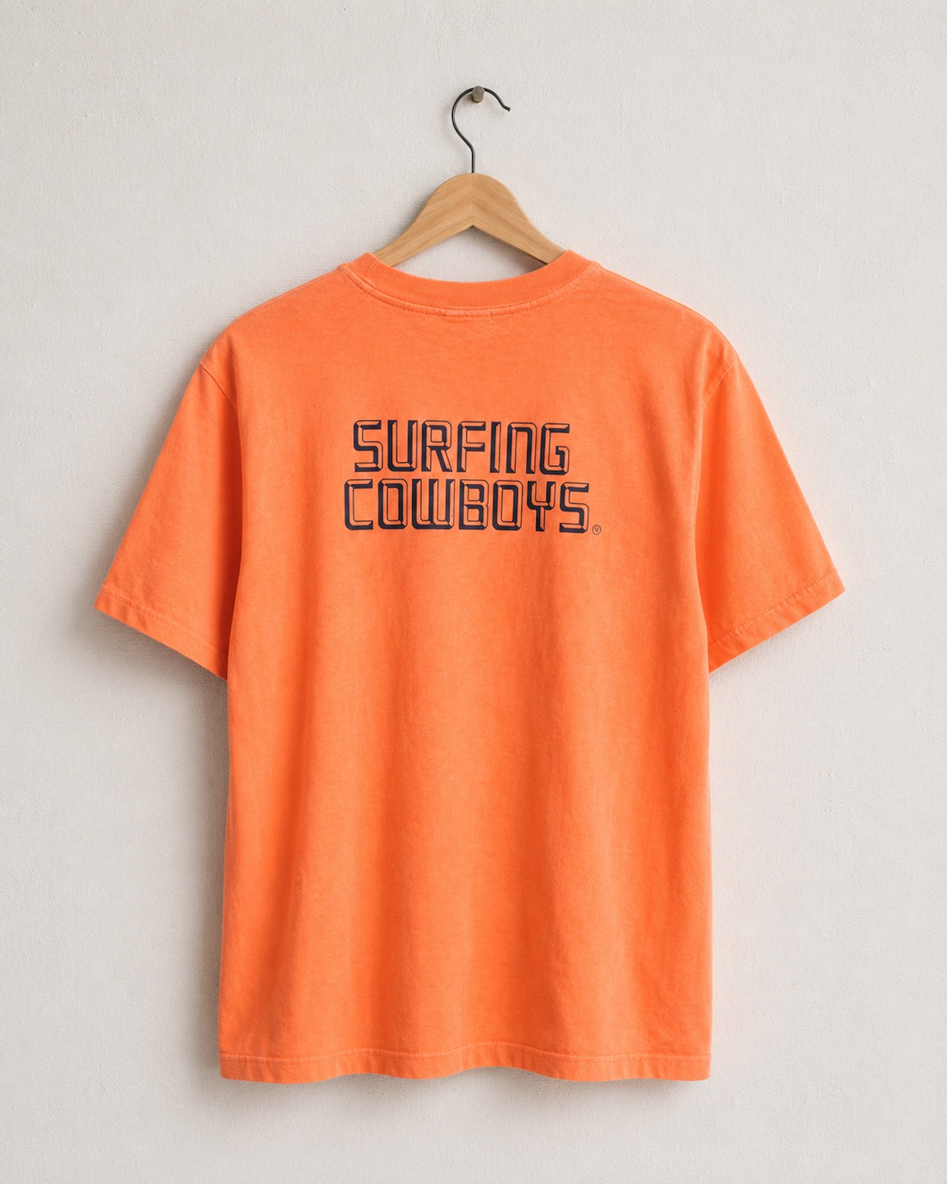 Faded orange t-shirt with 'Surfing Cowboys' text on a hanger against a light gray background