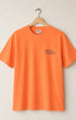 Faded orange t-shirt with 'Surfing Cowboys' logo on a hanger against a beige background