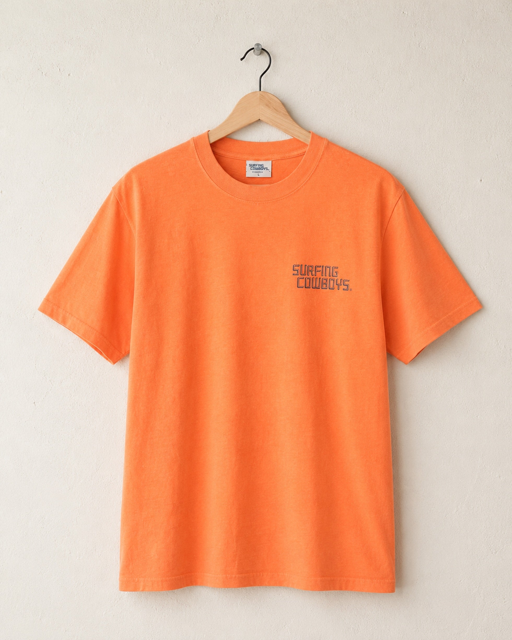 Faded orange t-shirt with 'Surfing Cowboys' logo on a hanger against a beige background