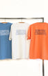 Three t-shirts in blue, white, and orange with 'Surfing Cowboys' text on a rack.