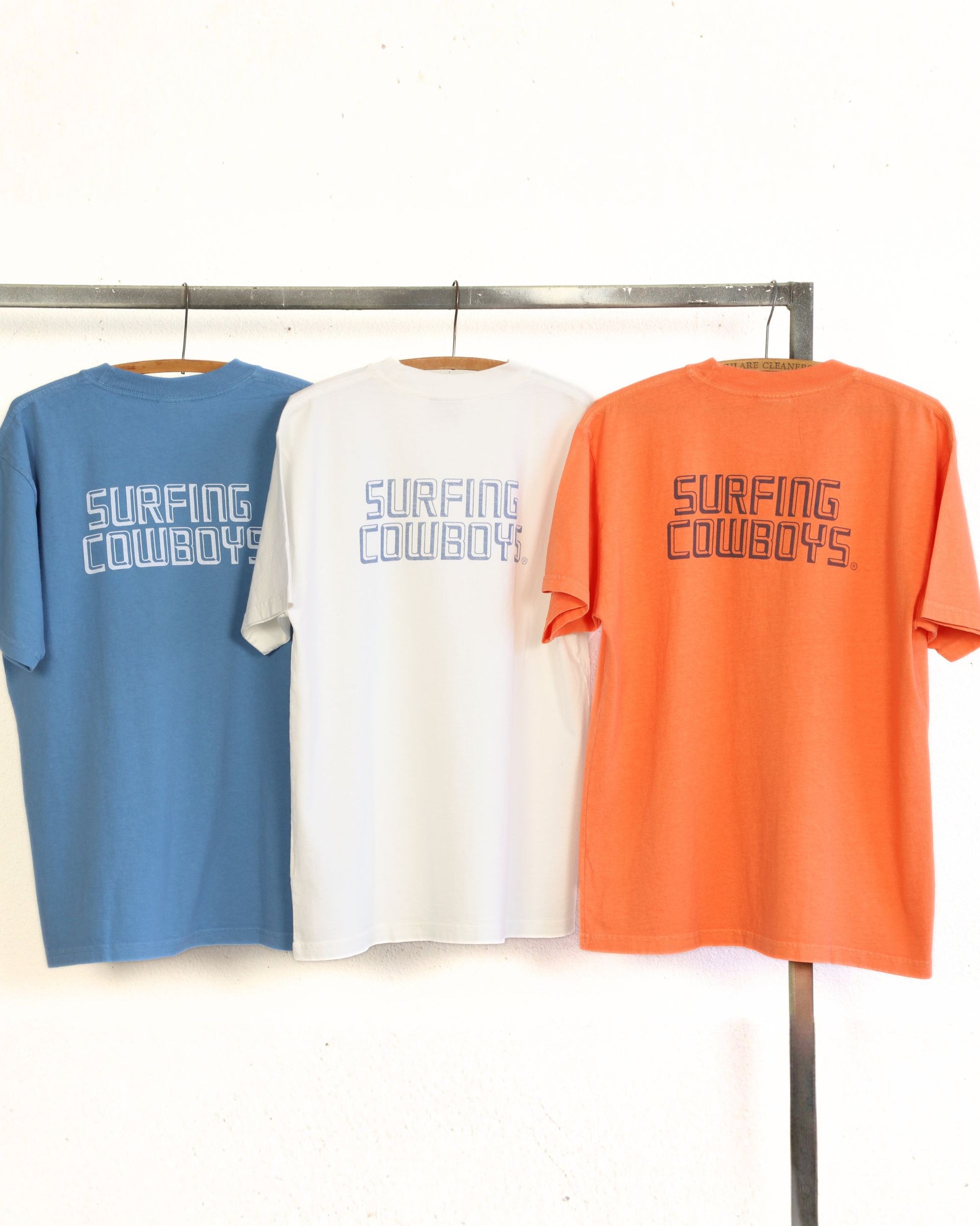 Three t-shirts in blue, white, and orange with 'Surfing Cowboys' text on a rack.