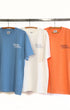 Three t-shirts in blue, white, and orange hanging on a rack with 'Surfing Cowboys' branding.