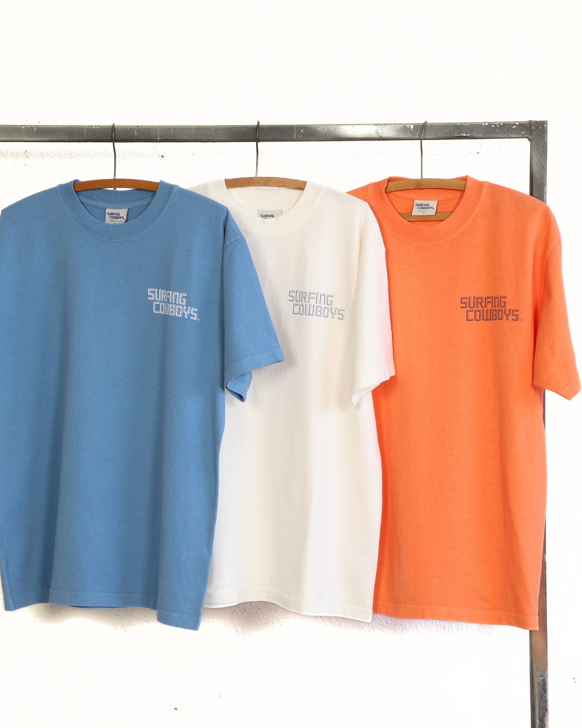 Three t-shirts in blue, white, and orange hanging on a rack with 'Surfing Cowboys' branding.