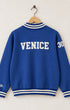 Back view of the blue half-zip sweatshirt showing a bold cream block font reading VENICE arched across the upper shoulders with custom-milled striped knit bands at the collar and waist.

