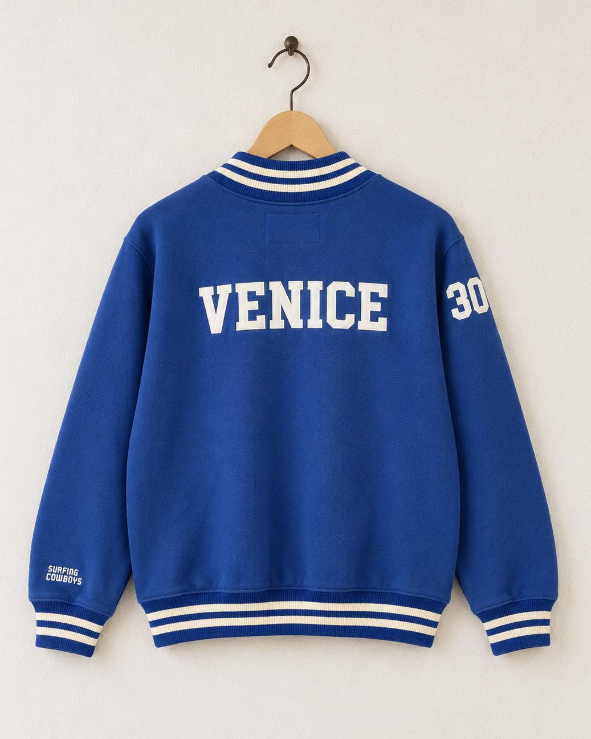 Back view of the blue half-zip sweatshirt showing a bold cream block font reading VENICE arched across the upper shoulders with custom-milled striped knit bands at the collar and waist.
