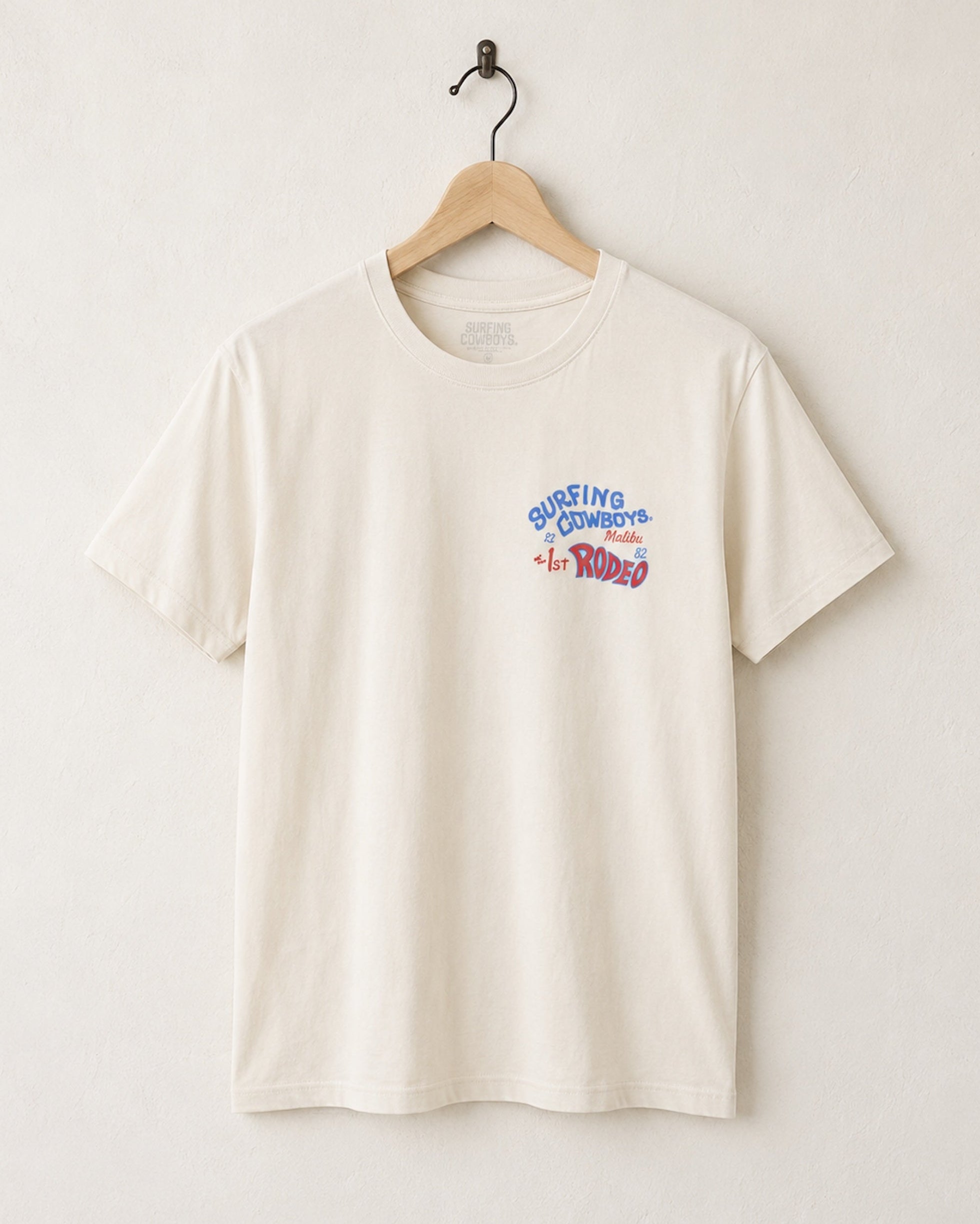 Front of Surfing Cowboys Rodeo cream colored t-shirt with chest placement colorful graphic on a hanger against a white background