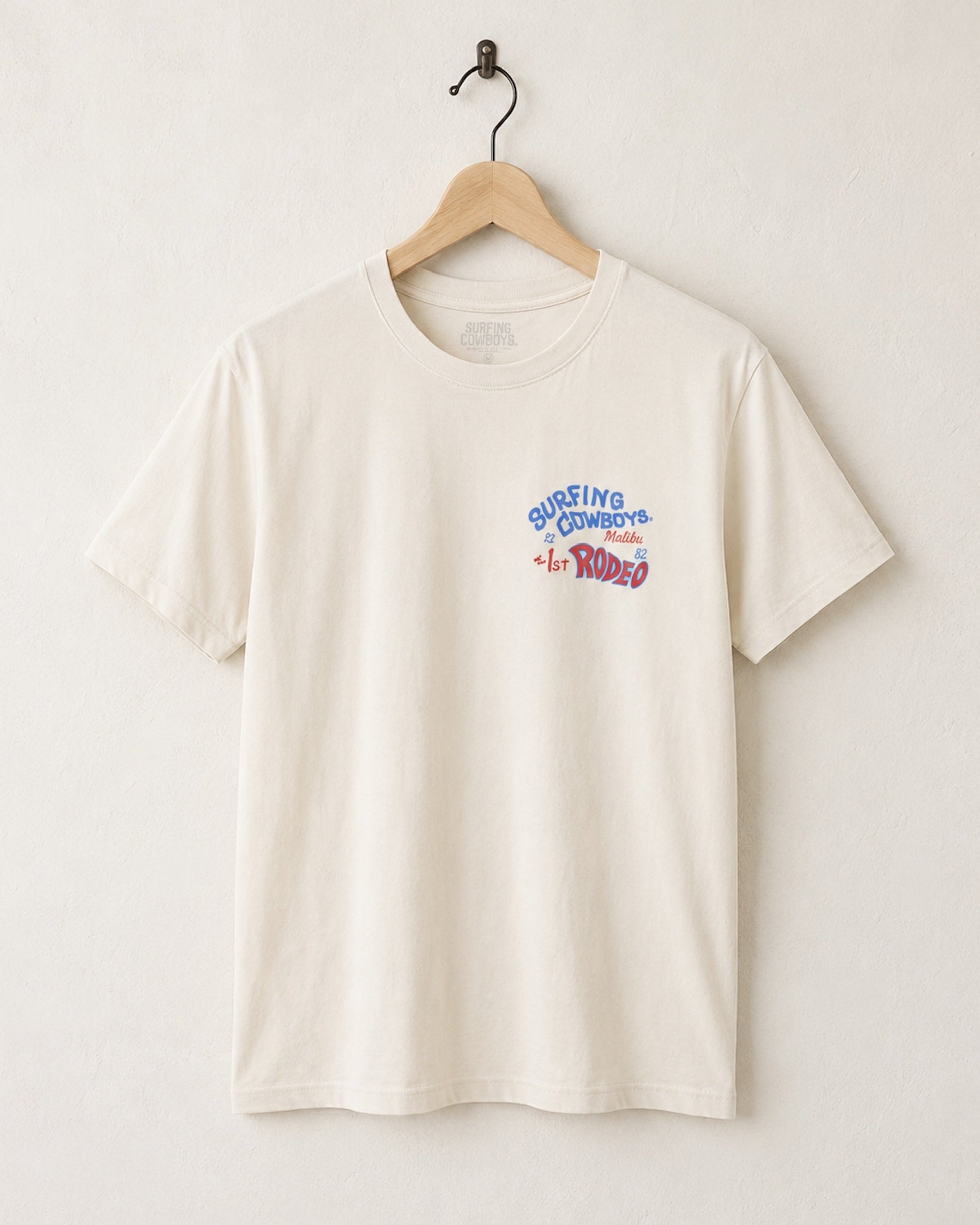 Front of Surfing Cowboys Rodeo cream colored t-shirt with chest placement colorful graphic on a hanger against a white background