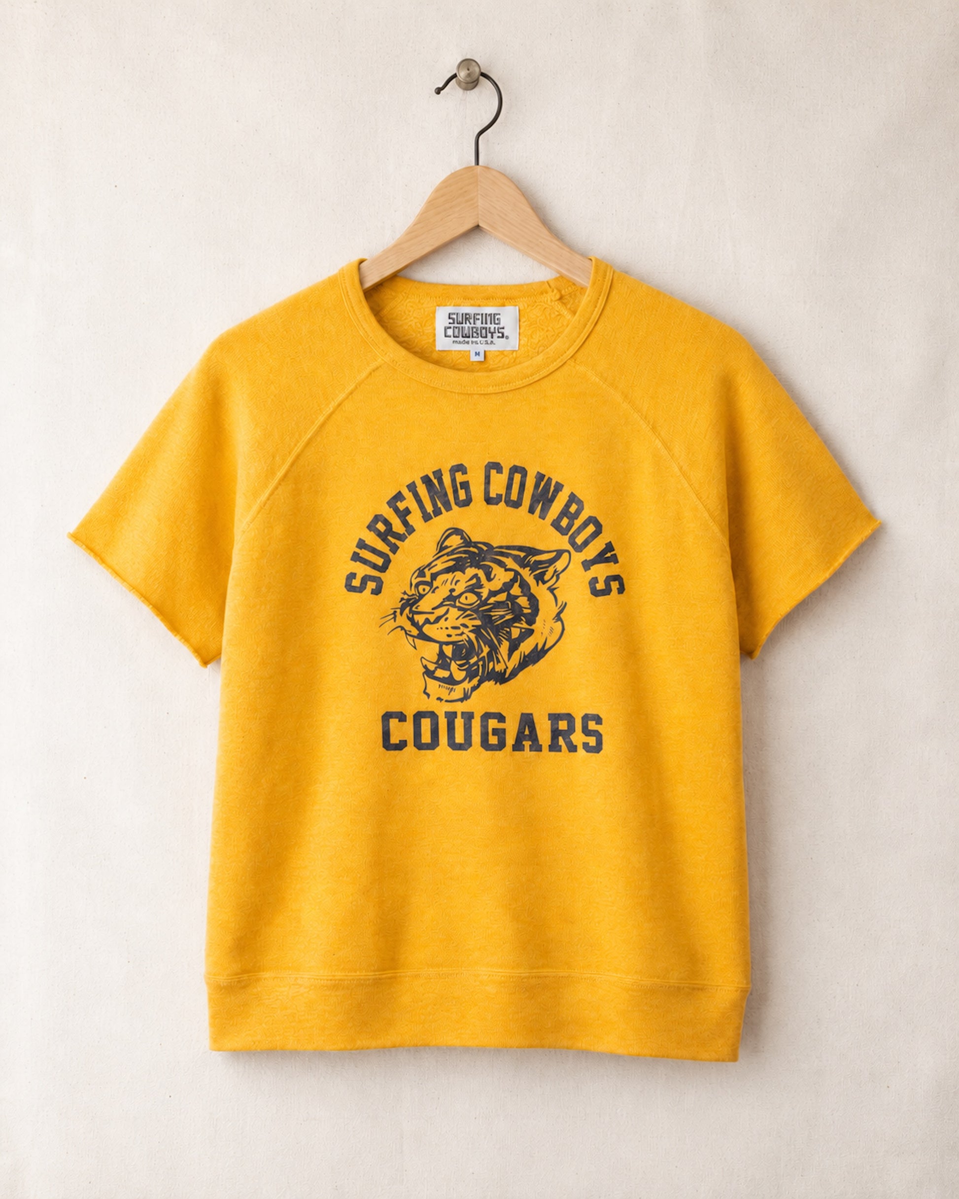 Yellow sweatshirt with 'Surfing Cowboys Cougars' text and tiger graphic on a hanger against a beige background