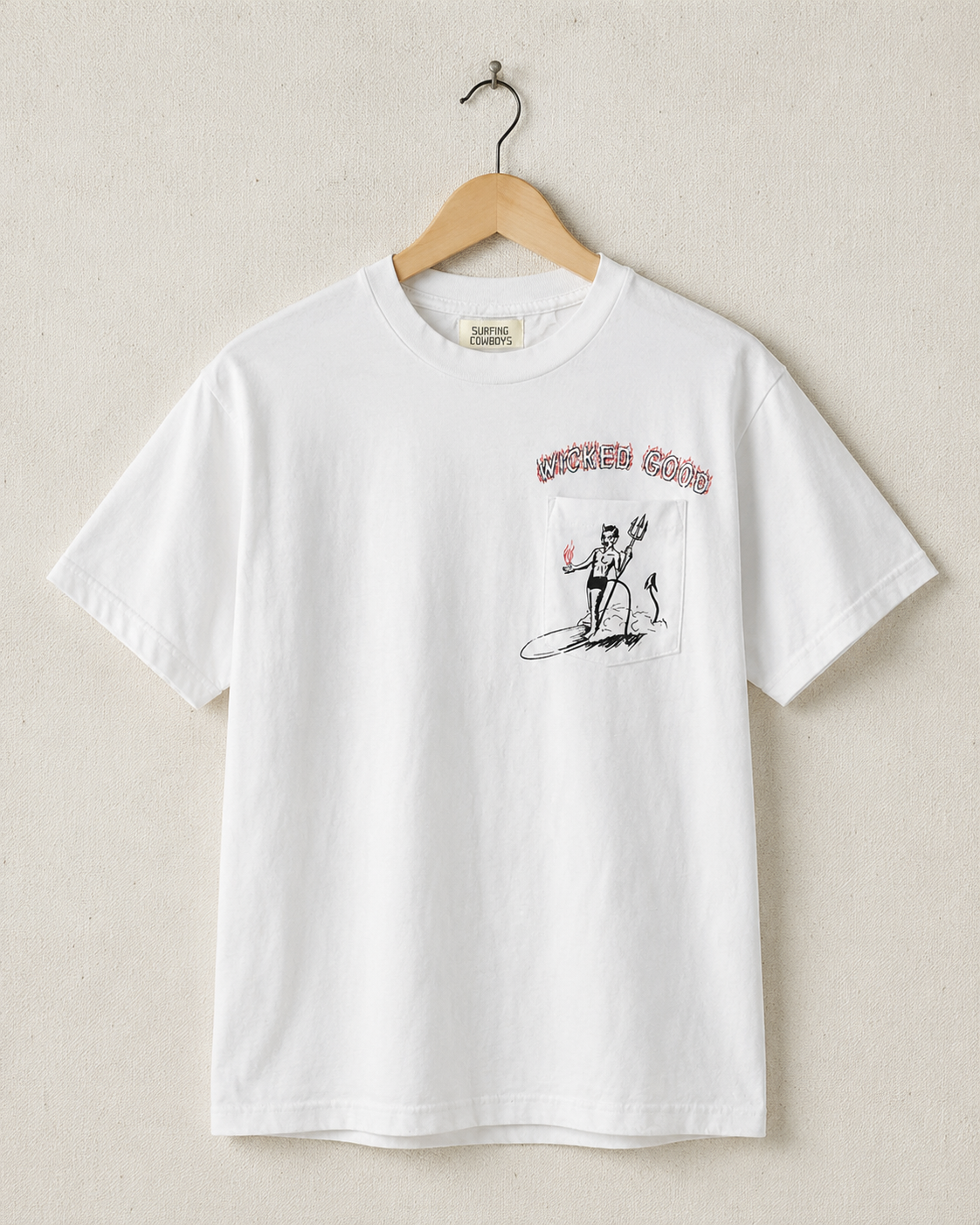 Front view of vintage white Surfing Cowboys Wicked Good pocket T-shirt with front devil graphic on hanger against tan wall. 