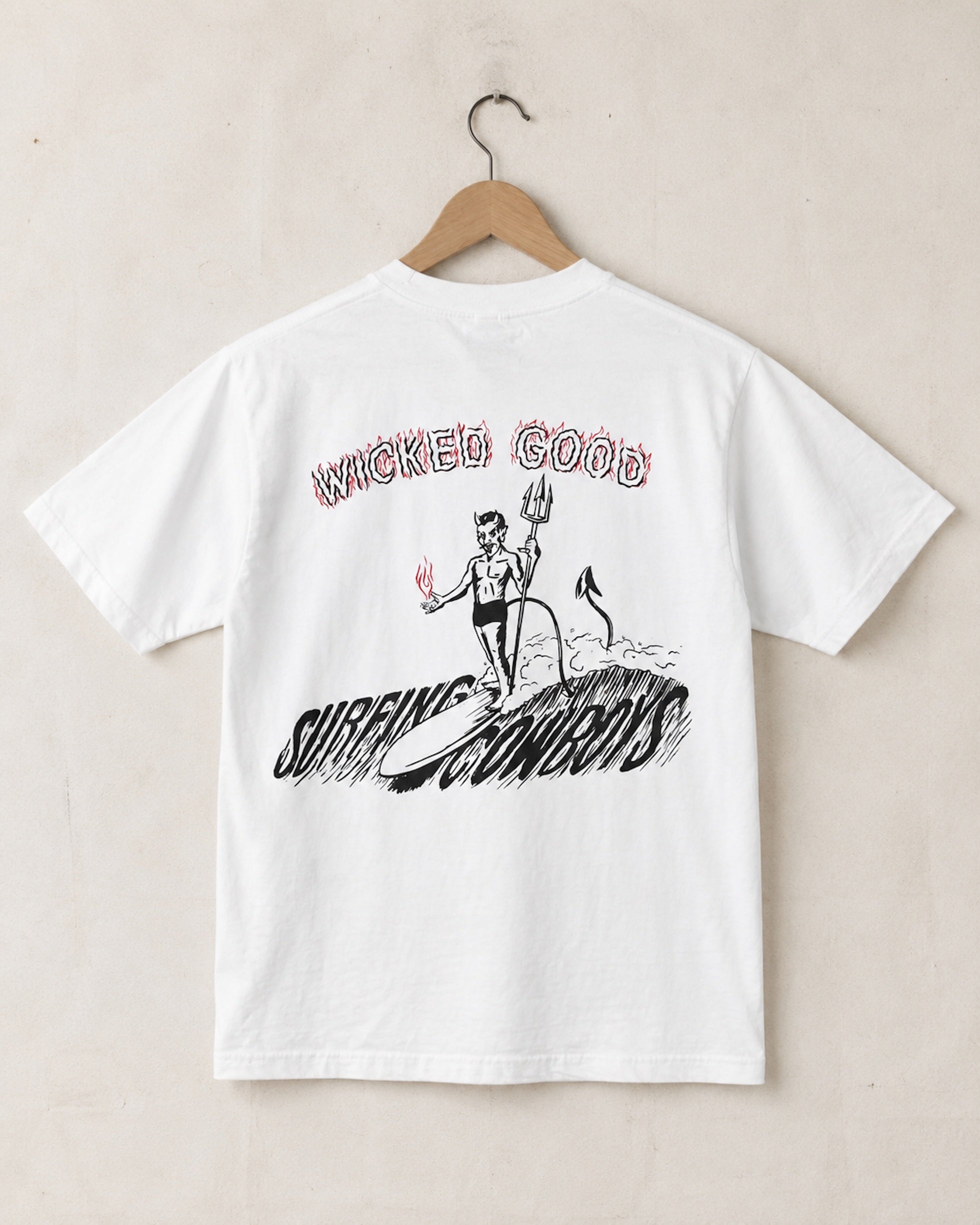 Back view of vintage white Wicked Good T-shirt with back devil graphic surfing a wave with Surfing Cowboys written in the water and Wicked Good written in the sky on hanger against tan wall. 