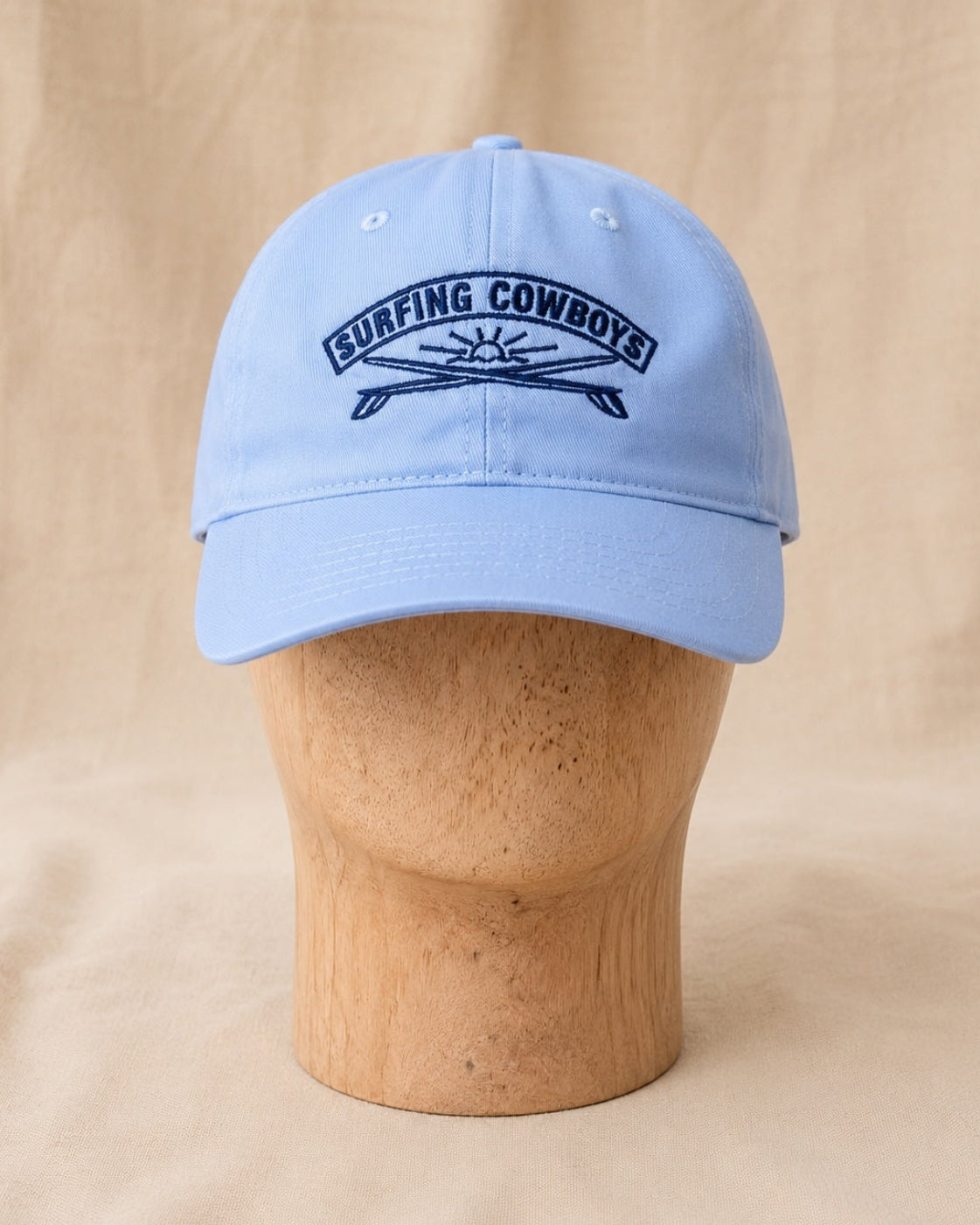 Front view of Surfing Cowboys Rising Sun Cap in light blue with navy embroidered crest on a wooden display head.
