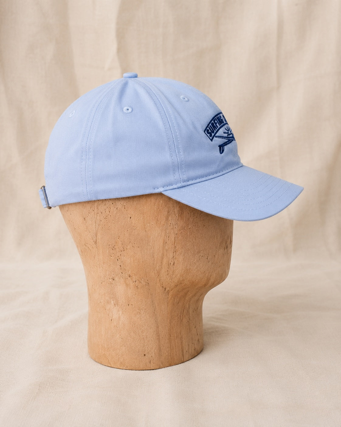 Side view of light blue Surfing Cowboys Rising Sun Cap showing curved bill, navy embroidered crest, and adjustable back strap.