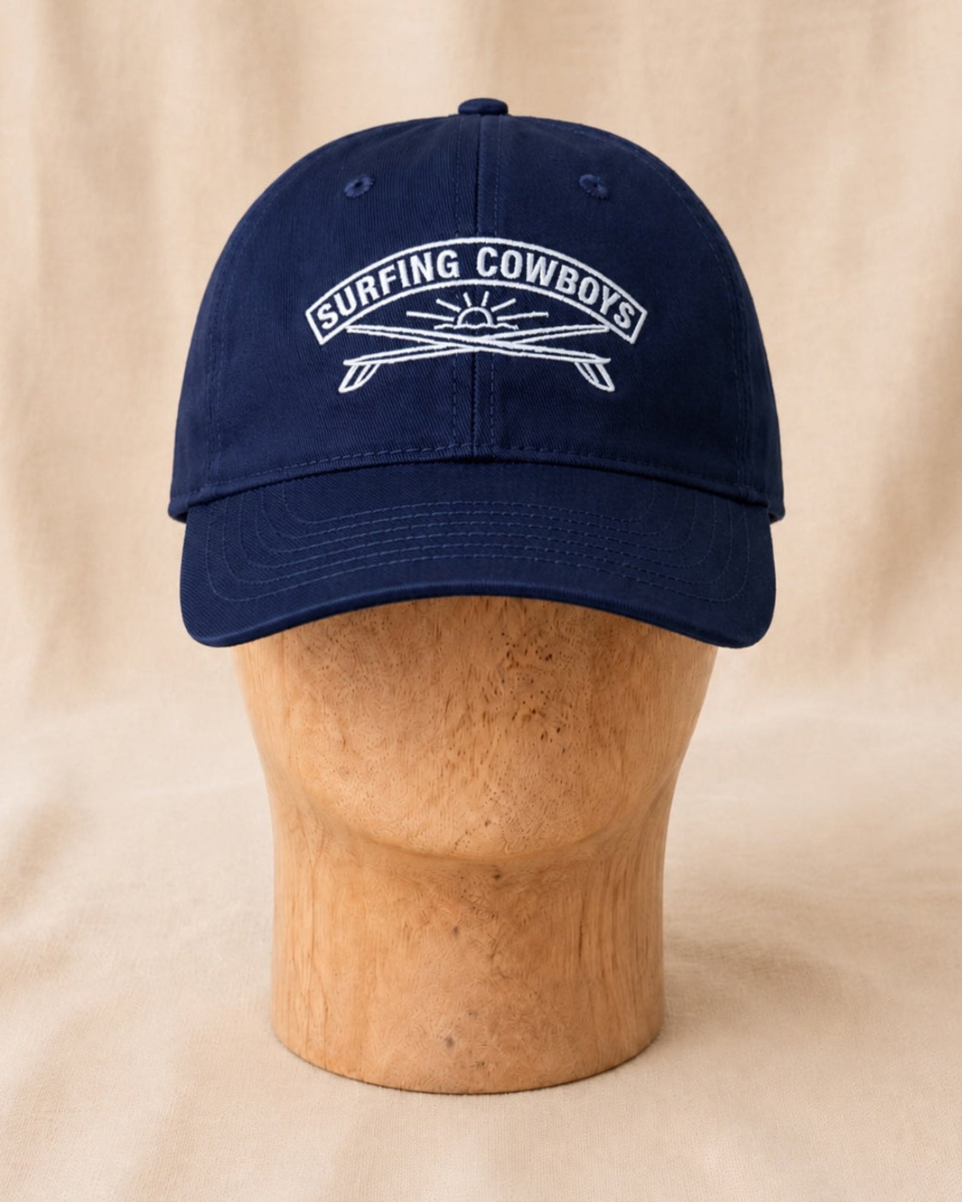 Front view of Surfing Cowboys Rising Sun Cap in navy with creamy-white embroidered crest on a wooden display head.